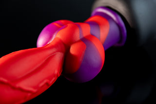 Close-up of the Fuxi fantasy dildo showing the red shaft, raised vein details, large rounded purple-and-red knot, and matte silicone texture on a dark reflective surface.